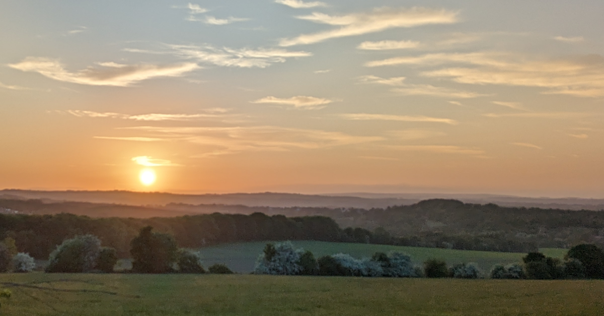 A picture of a sunset with hills and trees. There is mist in the trees below the sunset.