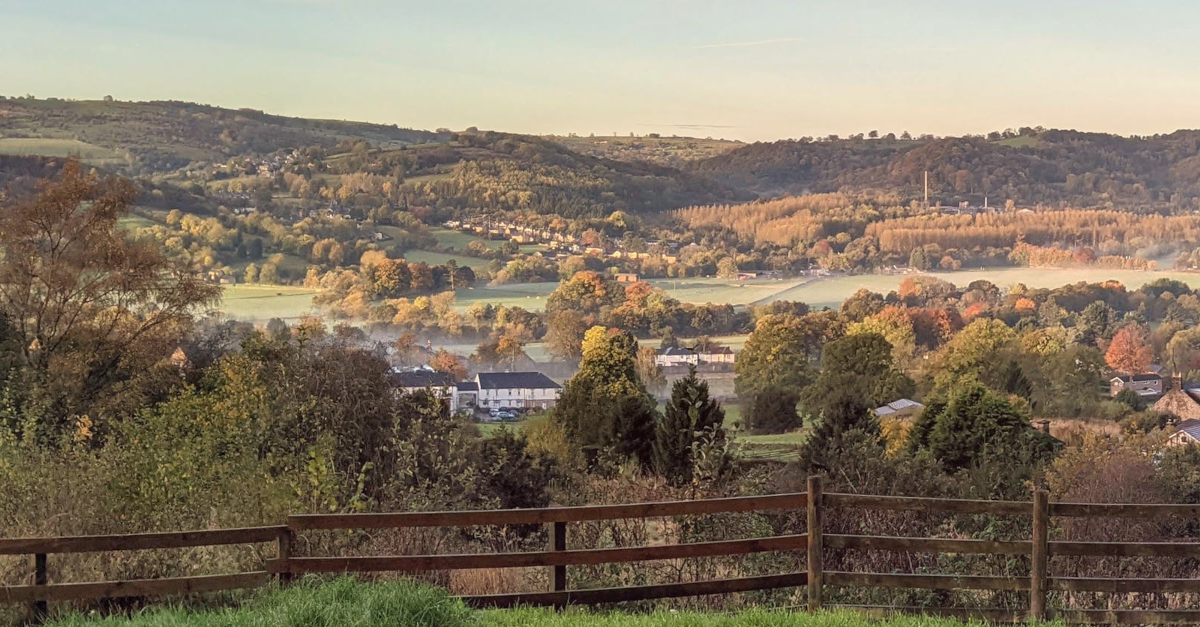 A scenic countryside landscape with rolling green hills, clusters of trees in autumn colors, and a small village nestled in the valley. A light mist lingers near the houses, while the morning or evening sunlight casts a warm golden glow across the fields. A wooden fence is visible in the foreground, framing the view.